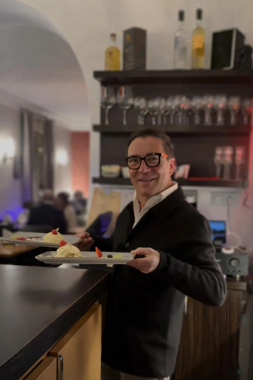 Der sympathische Gastgeber vom fiktiven Restaurant Genuss präsentiert am Tresen lächelnd zwei Teller mit einem liebevoll angerichteten Dessert.