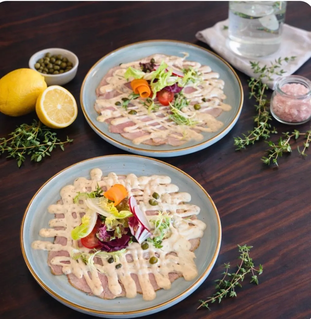 Zwei Portionen Vitello Tonnato mit frischer Garnitur und Beilagen auf einem rustikalen Holztisch.