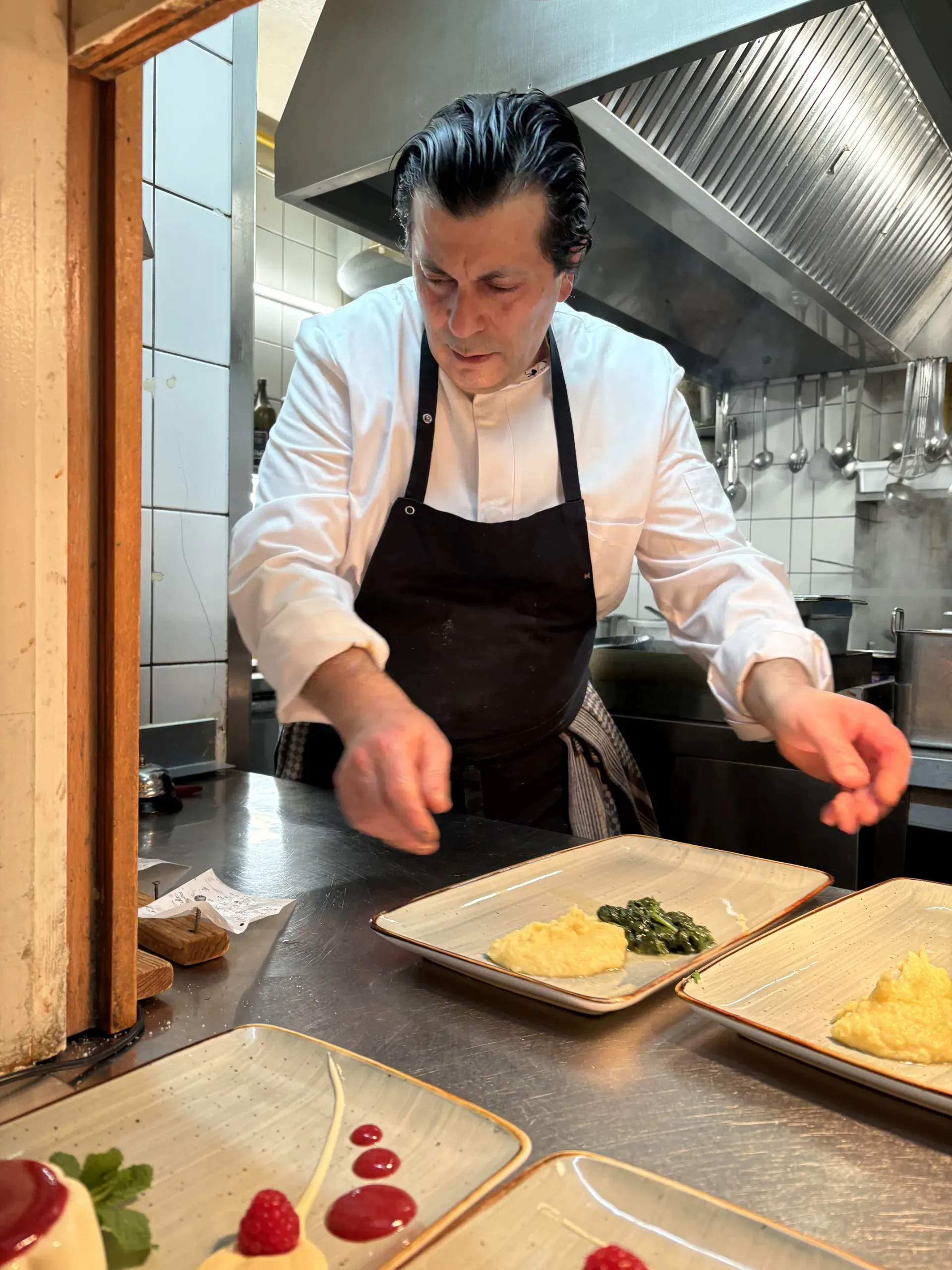 Ein Koch in weißer Jacke und schwarzer Schürze richtet in einer Restaurantküche konzentriert verschiedene Speisen auf rechteckigen Tellern an.